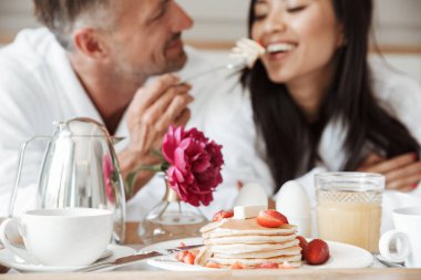 Mutlu genç çift kadın besleme adam yatakta yatarken romantik kahvaltı bornoz giymiş