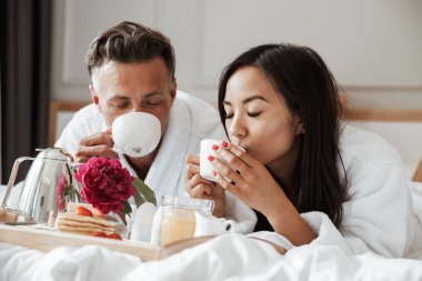 Çekici genç çift yatakta yatarken ve kahve içme sırasında romantik kahvaltı bornoz giymiş