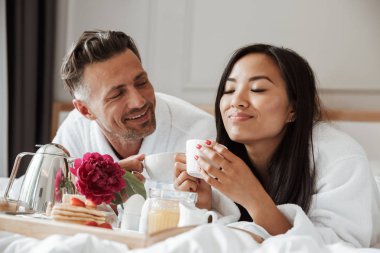 Yatakta yatarken ve kahve içme sırasında romantik kahvaltı bornoz giymiş güzel genç çift
