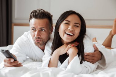 Çok ırklı kaç adam ve beyaz bornoz birlikte eve yatak odası ya da otel dairesinde uzaktan kumandayla el tv izlerken yalan giyen kadın fotoğrafı