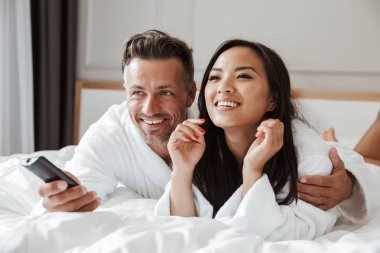 Çok ırklı çift beyaz adam ve uzaktan kumanda ile ev yatak odası ya da otel dairesinde el televizyon kanalları değiştirirken yalan beyaz bornoz giyen Asyalı kadın