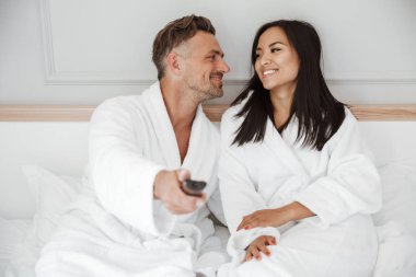 Güzel çift 30'lu yaşlarda beyaz adam ve beyaz elbisem gülümseyen ve uzaktan kumanda gösteriliyor ise ev yatak odası ya da otel dairede yalan giyen Asyalı kadın