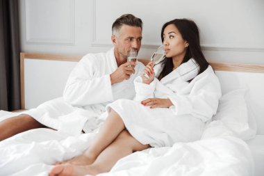 Mutlu güzel çift beyaz adam ve birbirlerine gülümsüyor ve otel odasında yatakta yalan süre gözlük köpüklü şarap içme Beyaz sabahlık giyen Asyalı kadın
