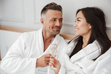 Romantik Çift 30'lu yaşlarda beyaz adam ve beyaz elbisem gülümseyen ve otel odasında yatakta yalan süre Gözlük şampanya içmek giyen Asyalı kadın