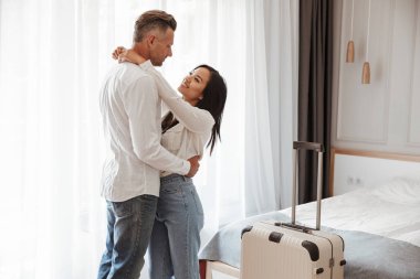 Görüntü çok ırklı çift erkek ve kadın casual giyim birlikte otel odasında büyük bagajlı sarılma giyiyor. Tatil kavramı