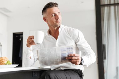 Adam gülümseyen bir mutfak ile bir gazete üzerinde otururken kahvaltı resmi elbise giymiş.