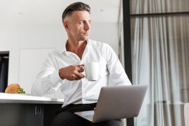 Kendine güvenen adam evde oturan ve kahvaltı ederken dizüstü kullanarak resmi elbise giymiş