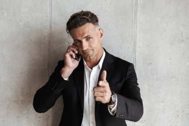 Cep telefonu üzerinde konuşurken ve gri duvar arka plan üzerinde parmak işaret takım elbise giymiş yakışıklı adam