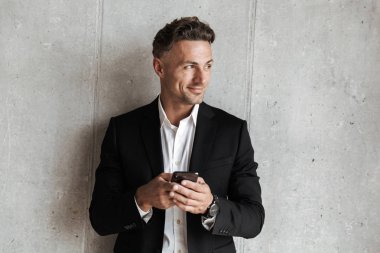 Yakışıklı adam cep telefonu tutan ve uzak gri duvar arka plan üzerinde seyir takım elbise giymiş