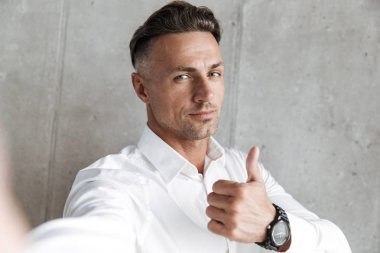 Beyaz gömlekli bir selfie outsretched el ile alarak ve başparmak gri duvar arka plan üzerinde gösterilen yakışıklı bir adam portresi