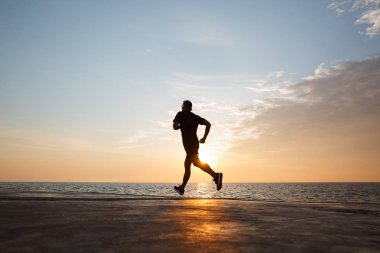 Atletik güçlü adam 30'lu şort ve tişört deniz ve gündoğumu sırasında kablosuz kulaklık ile müzik dinlemek rıhtımda boyunca çalışan silüeti