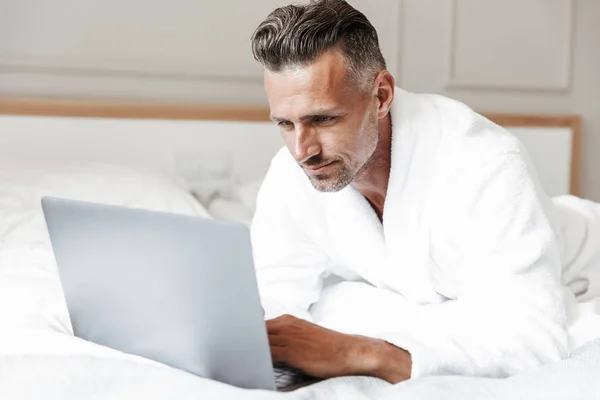Portrait of masculine adult man with gray beard wearing white housecoat looking at silver laptop while lying on bed in room or hotel apartment