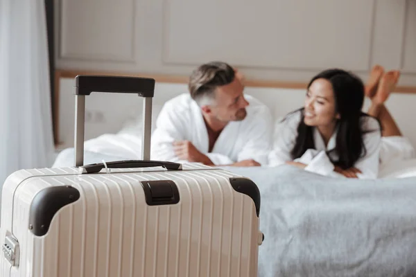 模糊多民族情侣的形象穿着白色浴衣躺在床上的大行李在酒店公寓的前景 度假理念 — 图库照片