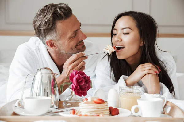 Genç çift gülümseyen kadın besleme adam yatakta yatarken romantik kahvaltı bornoz giymiş.