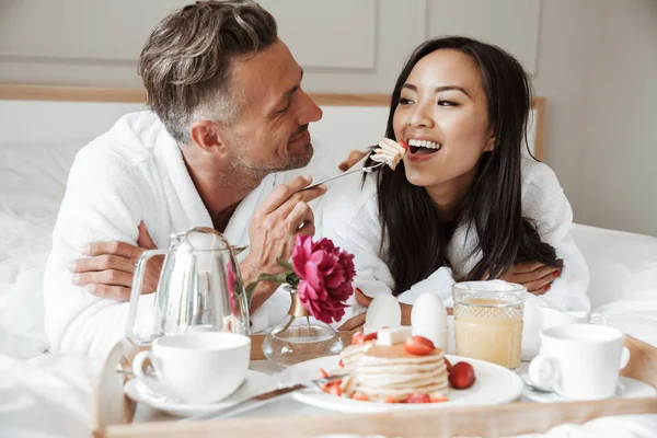 有吸引力的年轻夫妇穿着浴衣有浪漫的早餐 而躺在床上 男子喂养妇女 — 图库照片