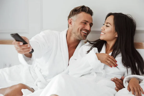 多民族夫妇白人男子和亚洲妇女穿着白色睡衣微笑着躺在家里的卧室或酒店公寓 而改变电视频道遥控器 — 图库照片