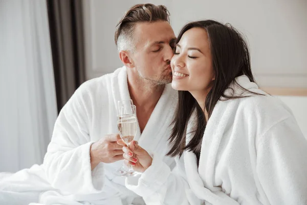 白人英俊男子的形象亲吻美丽的亚洲女人脸颊上 而喝香槟从眼镜在床上的酒店房间 — 图库照片