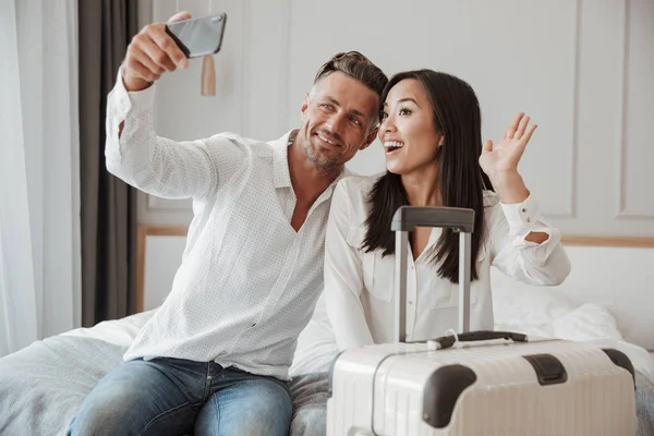 Mutlu genç çift alarak selfie otururken birlikte yatakta bir otel odasında bir bavulla cep telefonunuzla