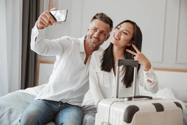 Genç çift alarak selfie otururken birlikte yatakta bir otel odasında bir bavulla cep telefonunuzla
