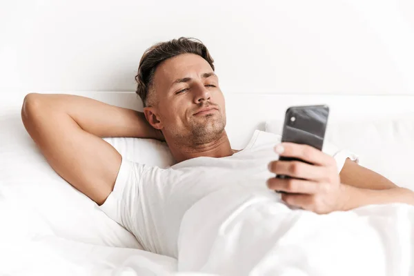 Yakışıklı Adam Yatakta Döşeme Sırasında Cep Telefonuyla — Stok fotoğraf
