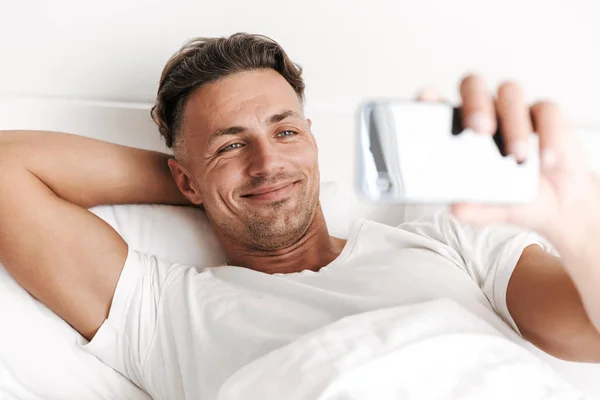 ベッドに敷設しながら携帯電話で Selfie を取って満足して男のクローズ アップ — ストック写真