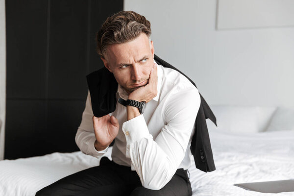 Уставший мужчина в костюме сидит на кровати и смотрит в сторону
