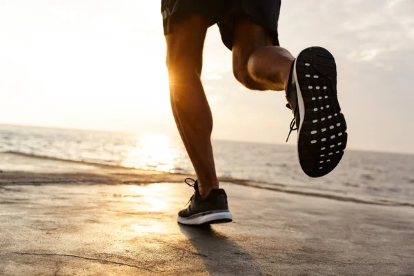 Cropped male legs of healthy sportsman wearing shorts and sneakers running along pier at seaside during beautiful sunrise