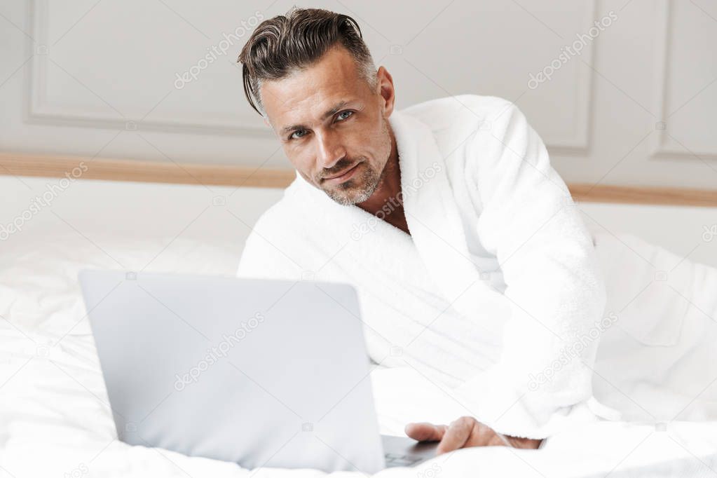 Image of european unshaved man with gray beard wearing white bathrobe looking at camera while lying in bedroom or hotel apartment with silver laptop