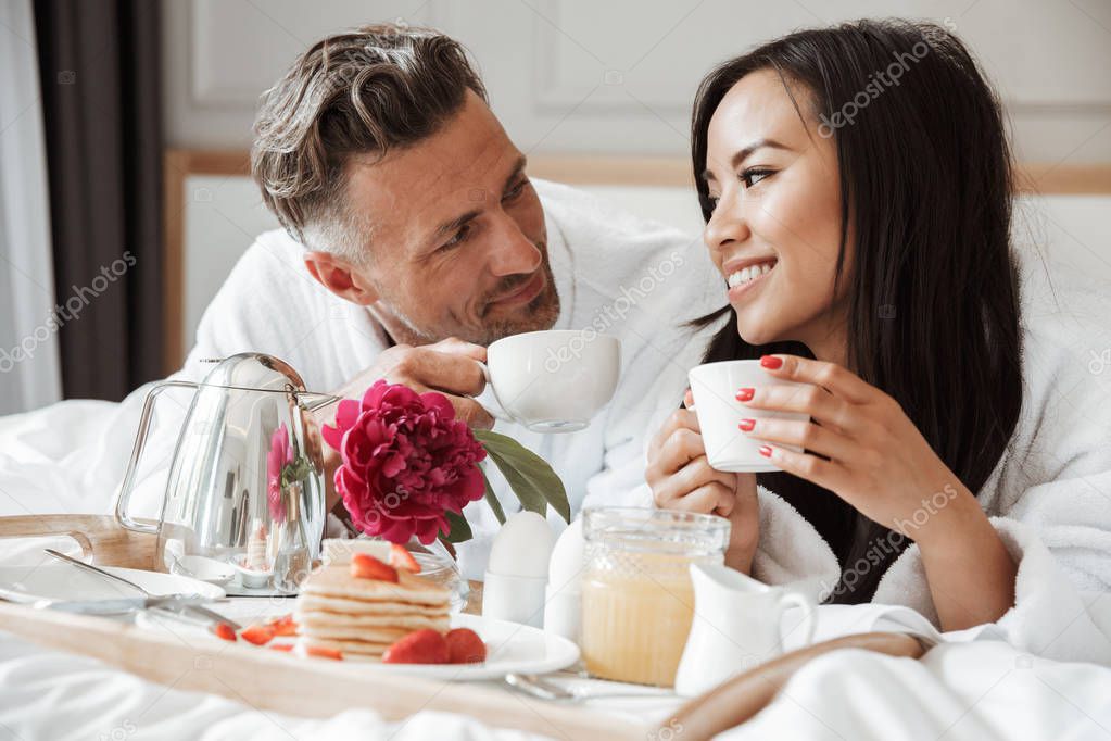 Smiling young couple dressed in bathrobes having romantic breakfast while lying on bed and drinking coffee
