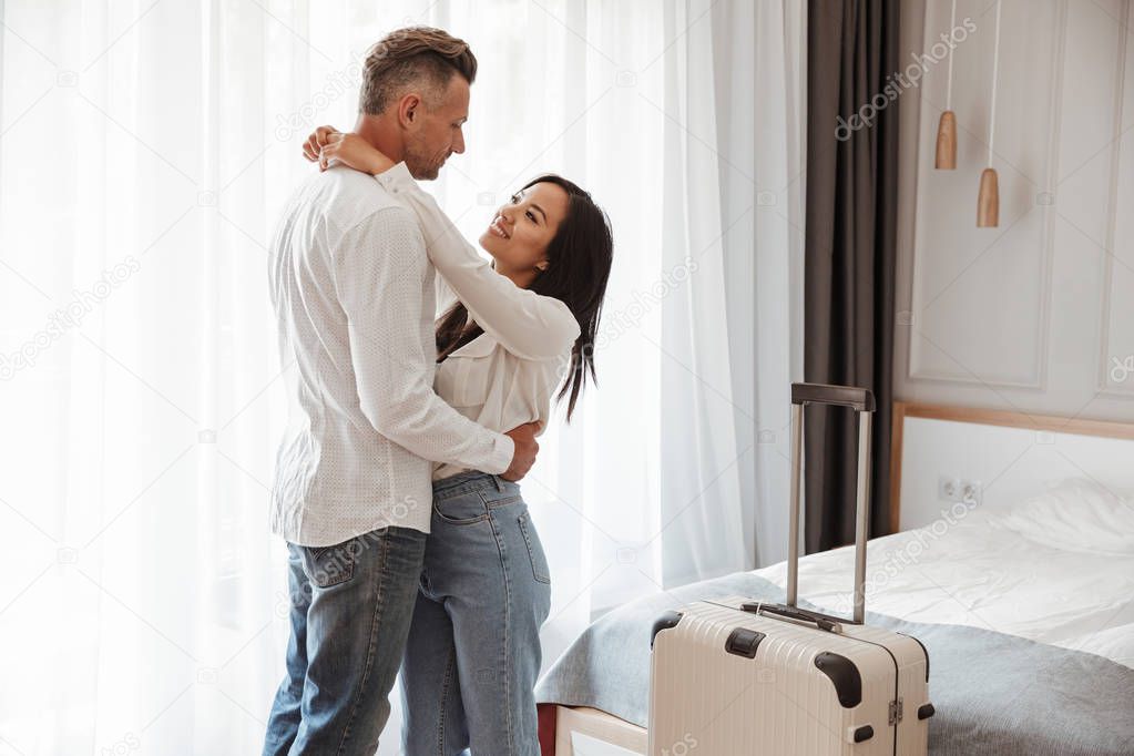 Image of multiethnic couple man and woman wearing casual clothing hugging together in hotel room with big luggage. Vacation concept