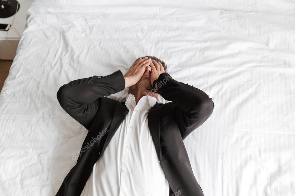 Tired man dressed in suit lying on bed with arms on his face