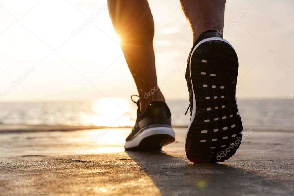 Image of sunlit caucasian athlete guy wearing sneakers running along pier at seaside during beautiful sunrise