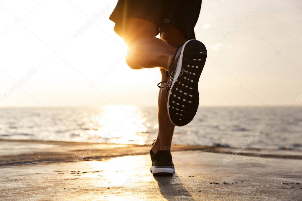 Cropped photo of active sporty man legs who wearing tracksuit and sneakers running along pier at seaside during beautiful sunrise