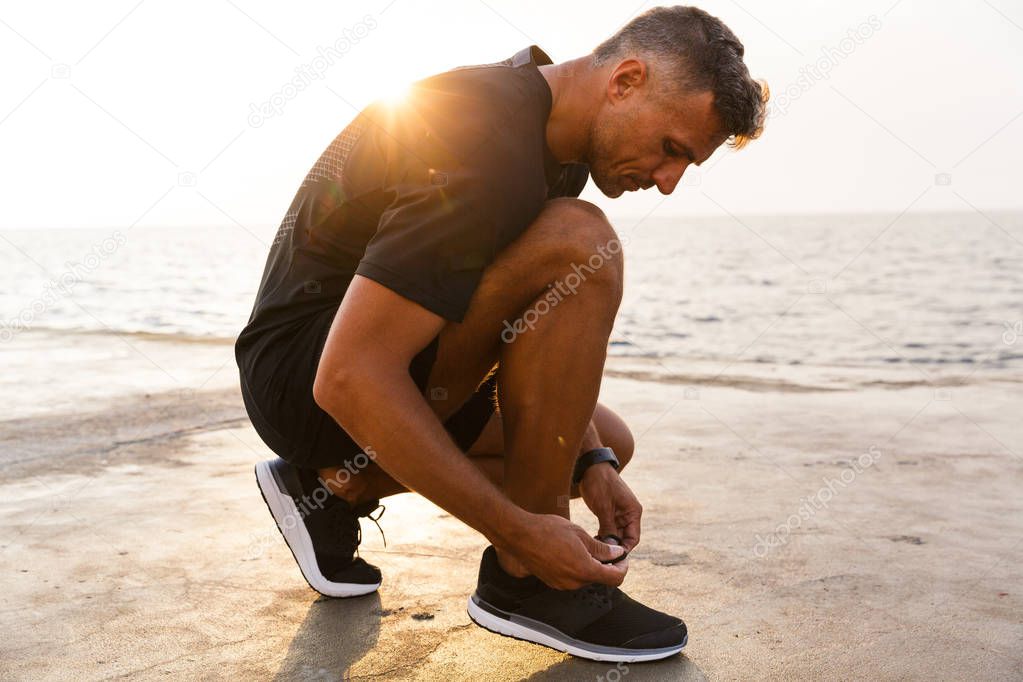 Photo of handsome caucasian man 30s in tracksuit and sneakers squatting on pier at seaside and tying his shoes