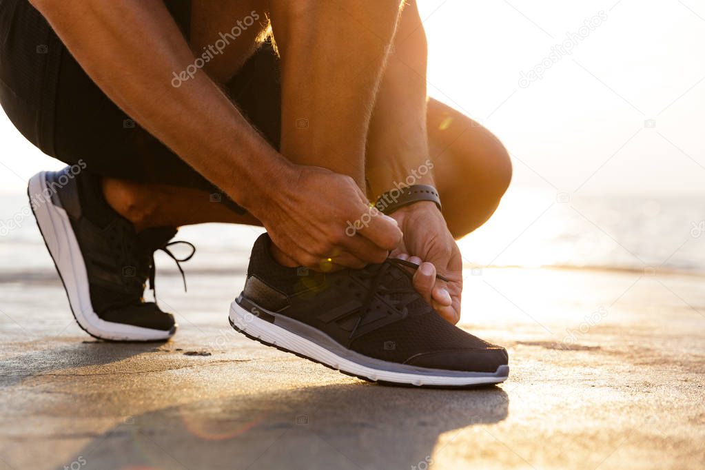 Cropped photo legs of sporty caucasian man in tracksuit and sneakers squatting on pier at seaside and tying his shoelaces