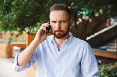 Karışık memnun Genç sakallı adam açık havada cep telefonuyla konuşurken fotoğrafı.