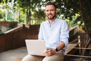 Fotoğraf gülümseyen duygusal Genç sakallı adam açık havada dizüstü bilgisayar kullanan.