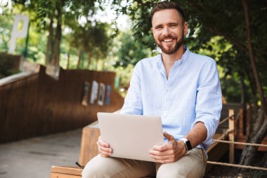 Fotoğraf gülümseyen duygusal Genç sakallı adam açık havada dizüstü bilgisayar kullanan.