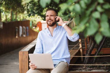 Fotoğraf gülümseyen duygusal Genç sakallı adam açık havada cep telefonuyla konuşurken dizüstü bilgisayar kullanan.