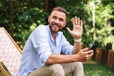 Fotoğraf gülümseyen duygusal Genç sakallı adam açık havada istimal hareket eden telefon bir yana sallayarak arıyorsunuz.