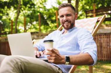 Fotoğraf gülümseyen duygusal Genç sakallı adam açık havada dizüstü bilgisayar kullanan.