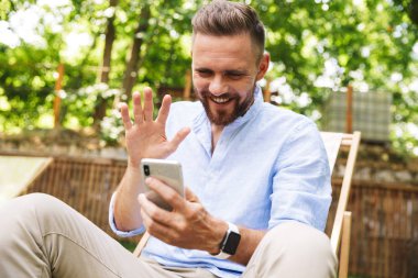 Mutlu genç sakallı adam açık havada cep telefonu sallayarak konuşuyor fotoğrafı.