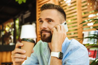 Mutlu sakallı yakışıklı genç bir kenara kahve içme cep telefonuyla konuşurken açık havada bakış fotoğraf.