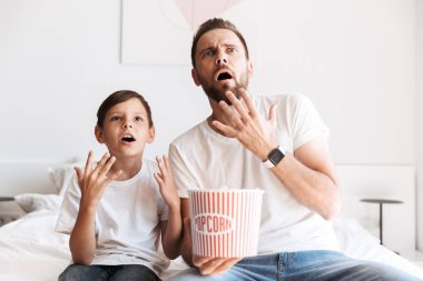 Şok genç adam baba baba ile oğlu evde kapalı pop mısır yemek Tv izlerken eğlenmek görüntüsünü.