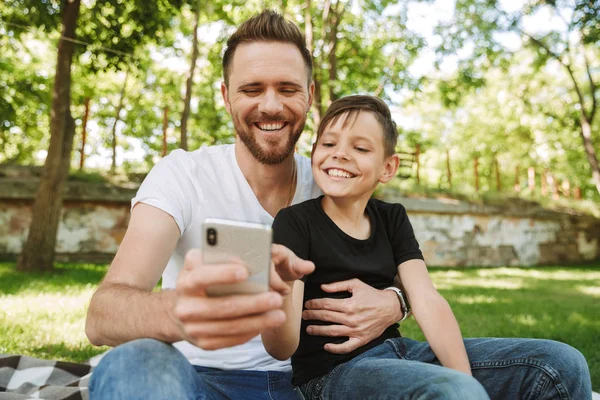 Mutlu genç baba oğlunun cep telefonunuzu kullanarak açık havada park doğa ile oturan görüntüsünü.