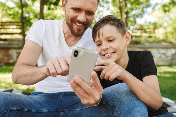 Görüntü bir cep telefonu kullanarak açık havada park doğa küçük oğluyla oturuyor neşeli genç baba.