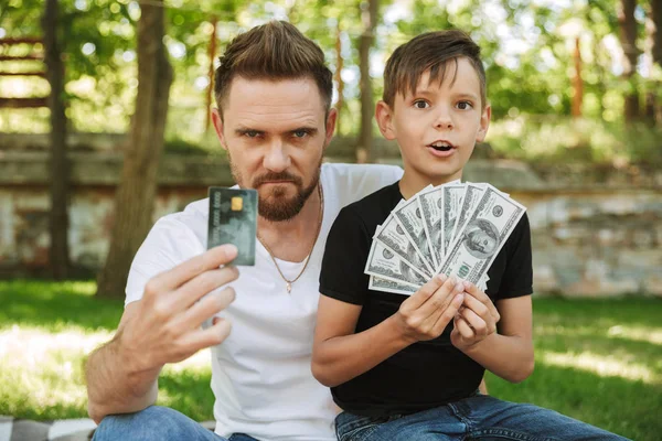 照片的严重年轻的父亲坐在户外公园自然与他的小儿子持有货币和信用卡 — 图库照片