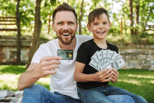 快乐兴奋的年轻父亲的照片坐在户外公园自然与他的小儿子持有货币和信用卡 — 图库照片
