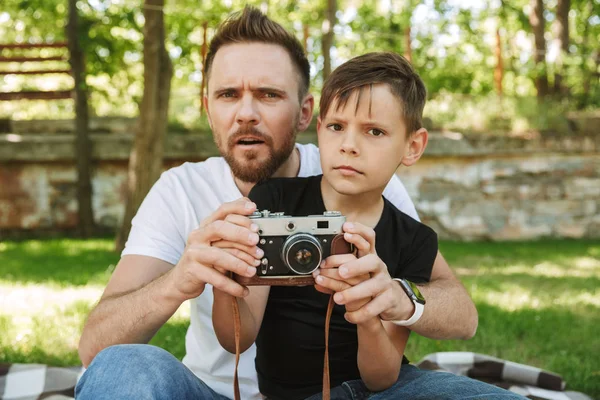 Görüntü bir açık havada park doğada kamera fotoğraf çekimi tutan küçük oğluyla oturan genç şaşkın baba.