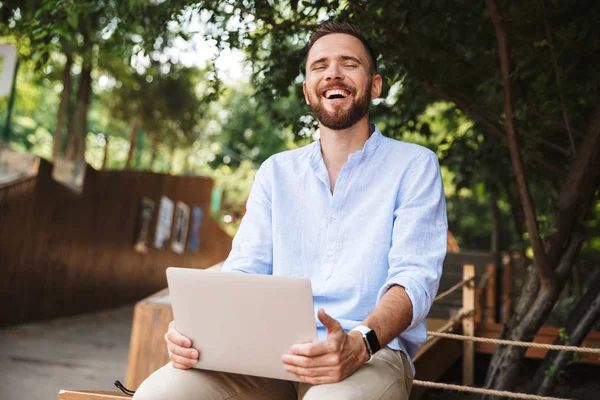 Fotoğraf gülümseyen duygusal Genç sakallı adam açık havada dizüstü bilgisayar kullanan.
